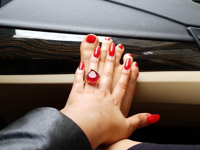 DomFrancaise's hand with shiny red nails, wearing a red gem ring, resting on a car's black and beige leather dashboard. Her ass is partially visible below.