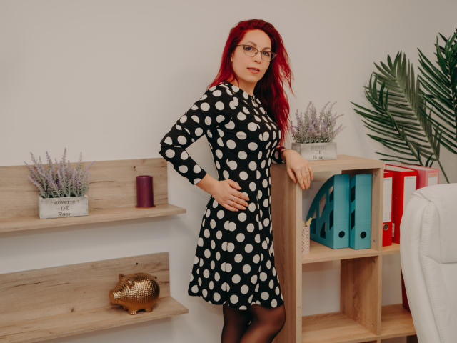 DirttyMature with red hair, glasses, black polka dot dress, black stockings, standing in room, hand on hip, wooden shelves with plants and colorful binders, white wall.