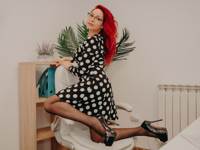 Een volwassen vrouw met rood haar, een bril, een zwarte jurk met stippen, zwarte kousen en zwarte hoge hakken, knielend op een witte stoel, kijkt over haar schouder, met haar billen omhoog..