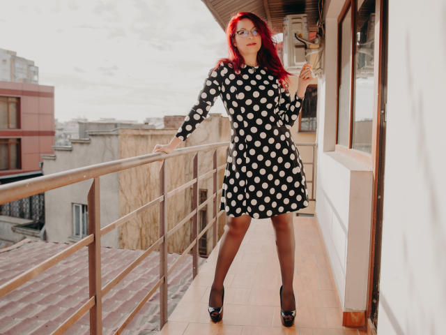 DirtyMature with red hair, glasses, black polka dot dress, black high heels, and black tights, standing on a balcony, holding a cigarette, looking confident.