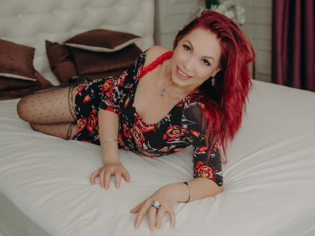 DirttyMature, avec des cheveux roux, portant une robe fleurie, un soutien-gorge en dentelle rouge et des collants à pois, allongée sur un lit, souriante, montrant ses seins, et portant une bague..