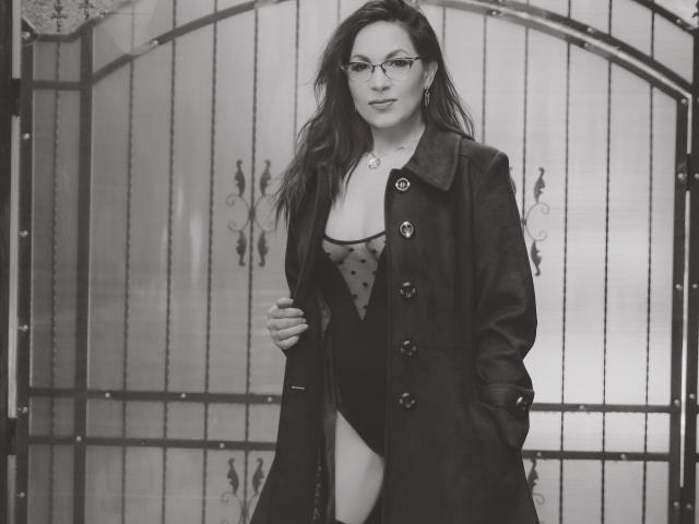 Black-and-white photo of DirttyMature with glasses, wearing a dark coat over a sheer, polka-dotted lingerie bodysuit, revealing her tits and a sliver of her ass.