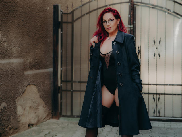 DirttyMature with red hair, glasses, black coat, and black lingerie showing her tits and pussy, standing in front of a metal gate. Her hands are in her coat pockets.