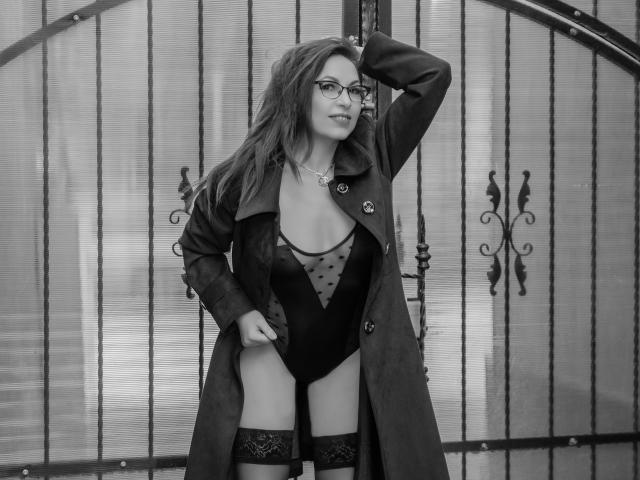 Black-and-white photo of DirttyMature with glasses, long dark hair, wearing a coat and black see-through bodysuit with polka dots, standing in front of a metal gate, showing her tits.