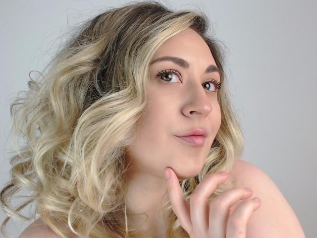 LuxNoir has blonde curly hair, green eyes, and is looking up with a slight smile, touching her chin with her right hand, showing her pink lips and subtle makeup.