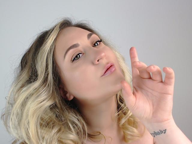 LuxNoir has blonde, wavy hair, a nose ring, and a tattoo on her right wrist, with her finger pointing up, looking at the camera with a slight smile.