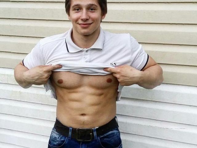 EdwinBull, a muscular young dude, lifts his white shirt to show off his defined abs and small, bare, pink nipples. He's wearing blue jeans with a black belt.