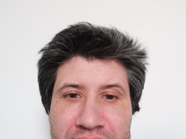 O rosto de MiraStud, com cabelo preto grisalho, levemente despenteado, olhos castanhos e um pequeno sorriso, com um fundo branco e um leve tom avermelhado no nariz..