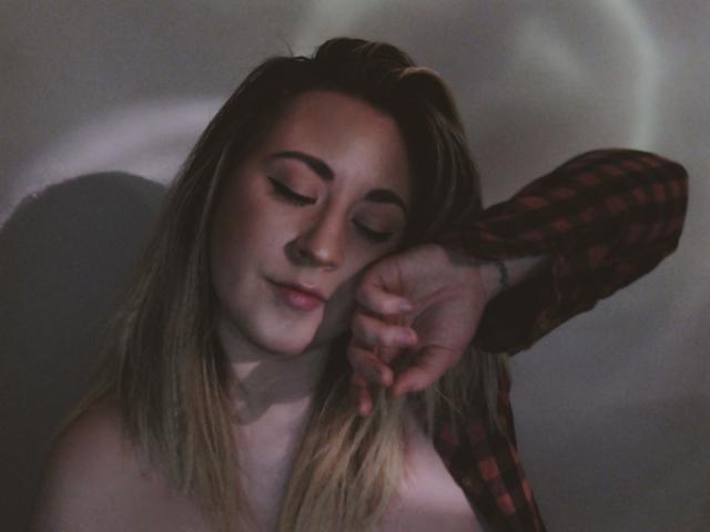 LuxNoir with blonde hair, eyes closed, wearing a red plaid shirt, touching her ear, soft smile, shadow on the wall, gentle light, bare shoulders, relaxed, subtle beauty.