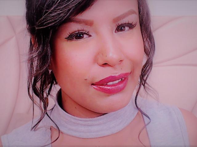 A ValleryC tem um rosto adorável, com cabelo castanho escuro, batom rosa e uma pequena perfuração no nariz. Ela está usando um colar cinza e olhando para a câmera com um sorriso..
