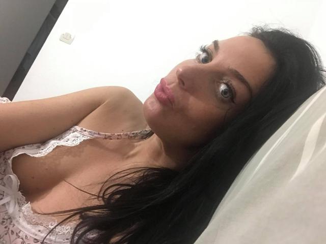 KatieFrenchie with pouty pink lips, dark wavy hair, and big blue eyes, wearing a white lace top, showing her small tits and slight cleavage in a selfie.