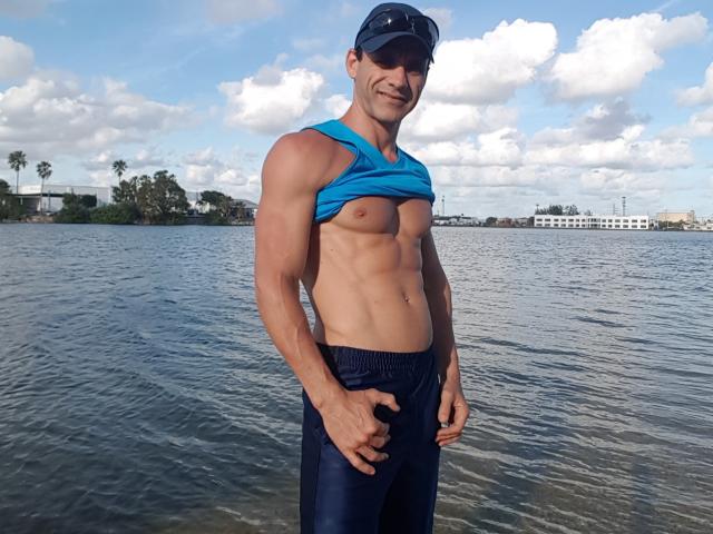EdenAdonis, sin camisa, gorra azul, musculoso, abdominales tonificados, pantalones azul oscuro, de pie en el agua, sonriendo, con gafas de sol, camiseta azul sin mangas, manos en los bolsillos, día soleado, cielo nublado..