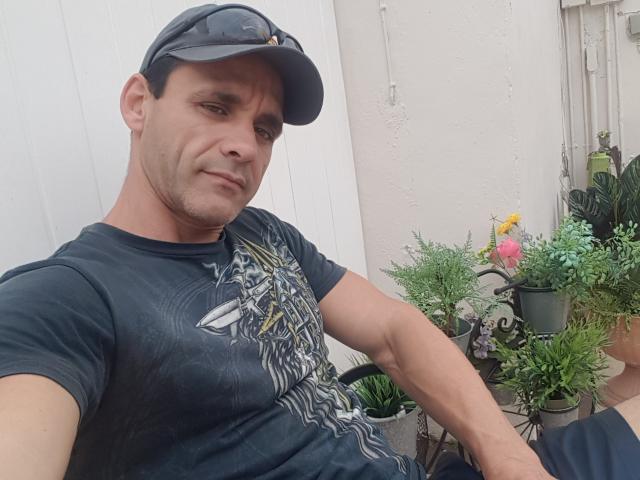 EdenAdonis, un hombre musculoso con una ligera sonrisa, que lleva una gorra de béisbol oscura y una camiseta con un diseño, está sentado con plantas en macetas al fondo. Su trasero es parcialmente visible..