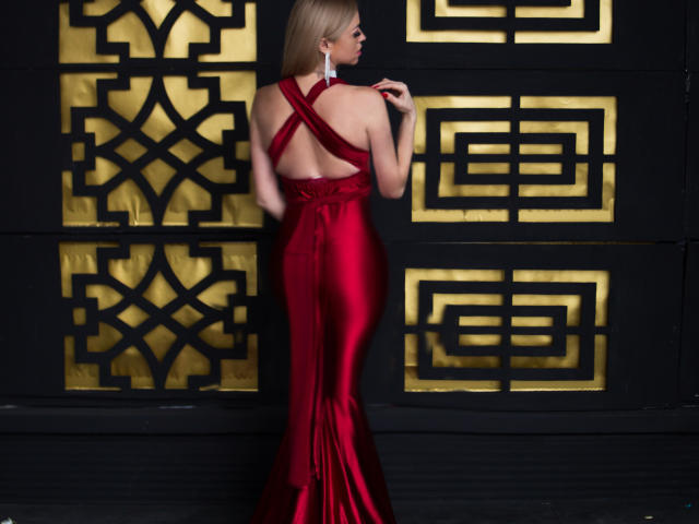 MiladyOne with long blonde hair in a shiny red satin dress, criss-cross back, standing against a black and gold geometric wall, showing her bare ass and elegant earrings.