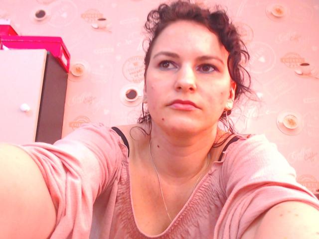 KinkyKosmina with curly brown hair, blue eyes, light skin, wearing a pink shirt, taking a selfie, showing a small mole on her chin, slightly parted lips, and a necklace.