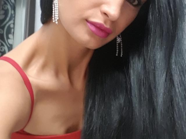 Het gezicht van TheSunshine, met roze lippenstift, zwart haar, een rode jurk en diamanten oorbellen, toont een slanke nek en schouderlijn. Borsten zijn gedeeltelijk zichtbaar, aan de linkerkant. Een zachte, sexy uitstraling..