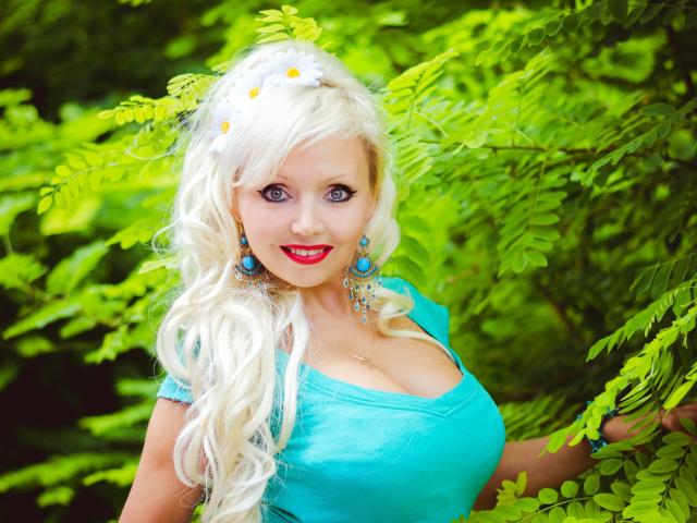 Blonde FurClubFetish with big tits, blue eyes, and red lipstick, wearing a turquoise top, blue earrings, and a white flower in her curly hair, standing among green leaves.