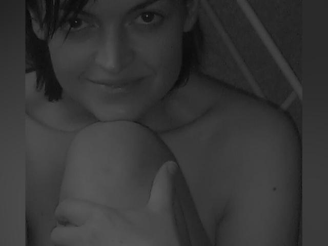 Grayscale photo of JulliGlei with wet hair, smiling slightly, holding a bald, circumcised penis. JulliGlei is nude, looking directly at the camera.