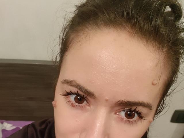 O rosto de GoldySquirt, com olhos castanhos, longos cílios e algumas pequenas espinhas na testa, com cabelo castanho desalinhado e uma expressão neutra, vestindo uma camisa escura..