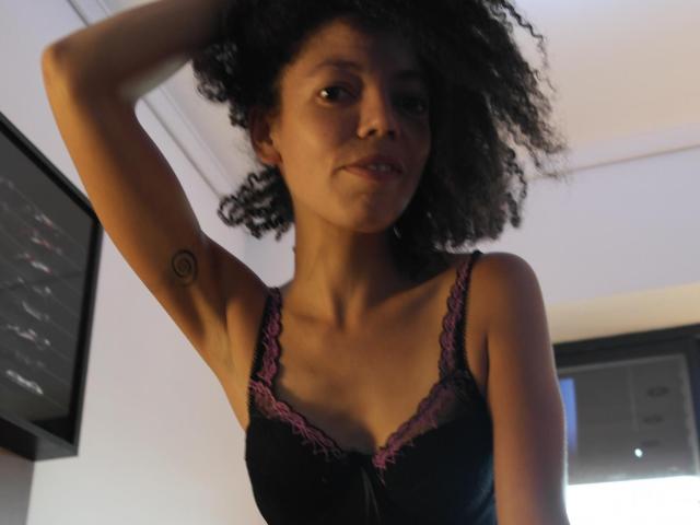 Una latina de aspecto soñador, con cabello negro rizado, lleva un sujetador negro con encaje y tiene un pequeño tatuaje circular en el brazo derecho. Está de pie con el brazo levantado, y sus senos son visibles..