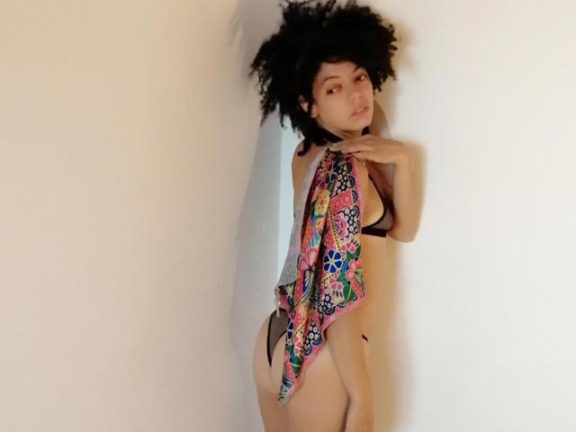 DreamyLatina with curly black hair, wearing a colorful floral one-piece, black thong, standing against a white wall, looking over shoulder, hands near neck, showing her ass.
