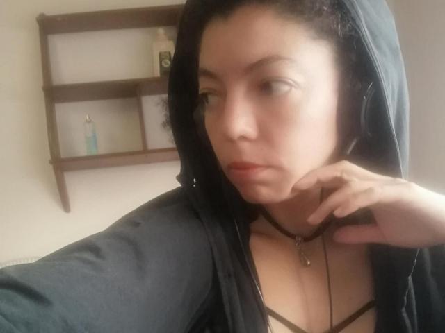 Latina con un aspetto sognante, pelle chiara, felpa nera con cappuccio e spalline del reggiseno marroni, che guarda di lato con la mano sul mento, indossando una collana con una piccola croce..