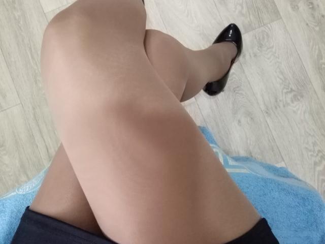 AnabelSex69 zit met gekruiste benen, draagt doorschijnende panty's en zwarte pumps van leer, en zit op een blauwe handdoek, met een houten vloer op de achtergrond..