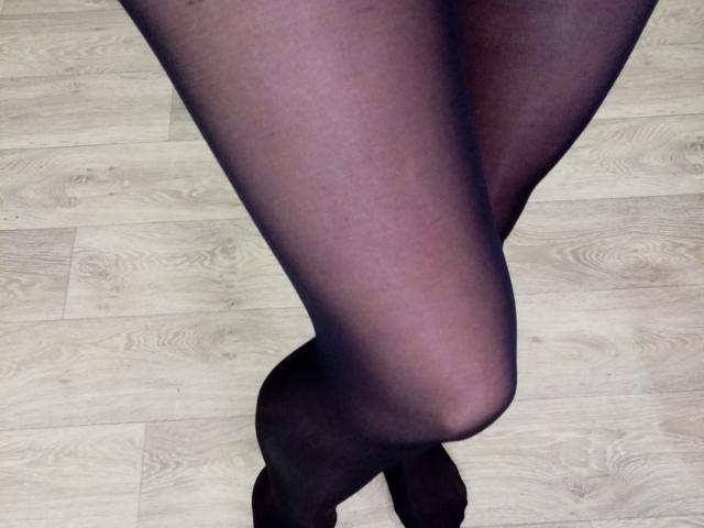 AnabelSex69s Beine in schwarzen, transparenten Strumpfhosen, die an den Knöcheln gekreuzt sind, zeigen glatte, dunkelbraune Haut und einen leichten Schatten auf einem hellen Holzboden..