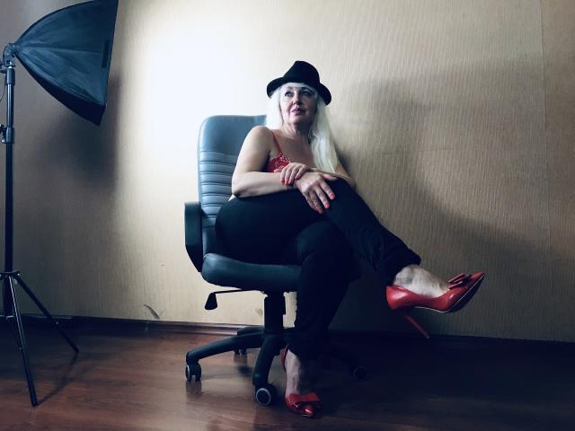 KamaliaSim with pale skin, long white hair, black hat, red lace top, black pants, red heels, sitting on a black chair, legs crossed, smiling, wooden floor, studio light.