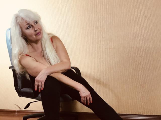KamaliaSim, com cabelo branco comprido, sutiã vermelho, calças legging pretas e unhas vermelhas, sentada em uma cadeira, pernas abertas, braços cruzados, olhando para a câmera com um leve sorriso..