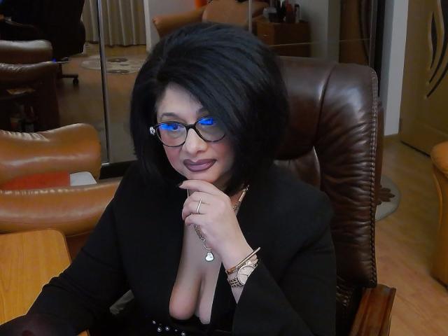 ClassybutNaughty, met zwart haar, een bril en een zwarte outfit, die haar decolleté laat zien met haar hand omhoog, met een ring en armband, zittend in een leren stoel..