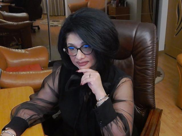 ClassybutNaughty, avec ses cheveux noirs, ses lunettes bleues et son haut noir transparent, assise dans un fauteuil en cuir marron, le menton appuyé sur sa main, montrant ses seins et une bague..