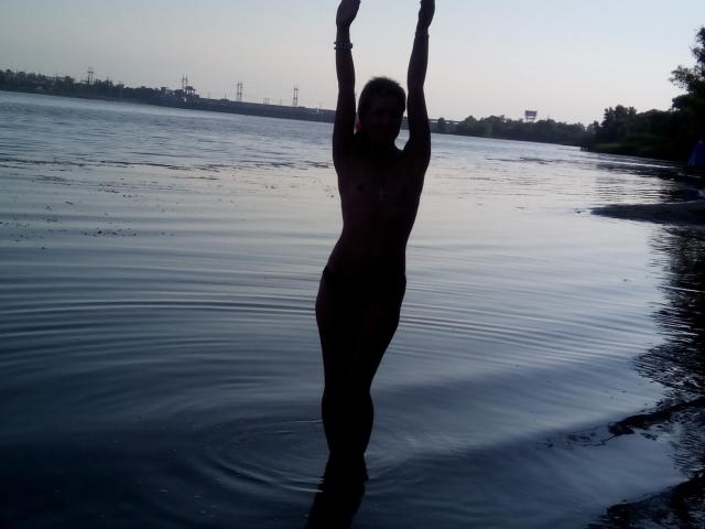 Silhouet van Alena. Blond, naakt, met armen omhoog, staand in het water, met een slank, getraind lichaam, kleine borsten en donkere huid, in de schemering, met rimpelingen rond haar benen..