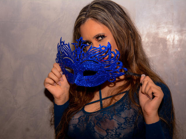 GiovannaSweet, com longos cabelos castanhos, vestindo uma blusa azul de renda, segura uma máscara de carnaval azul brilhante que cobre seus olhos, e suas mãos seguram as laterais da máscara..