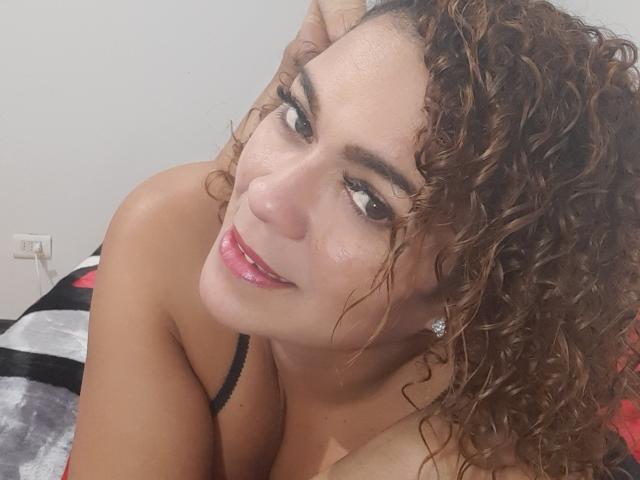 SolmaryHot tem cabelos castanhos cacheados, um lábio rosa brilhante e um leve sorriso. Ela está olhando para cima com olhos castanhos, usando uma alça preta. Seus seios estão parcialmente visíveis..