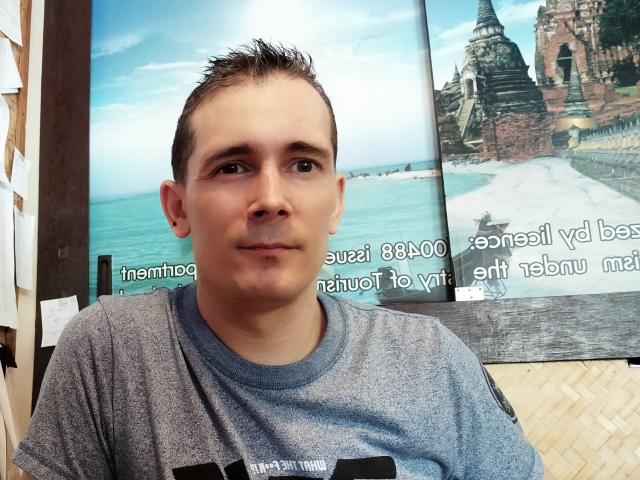 Kinkme, um rapaz de cabelo castanho curto, com um leve sorriso, vestindo uma camisa cinza, sentado em um lugar aconchegante, com uma foto de uma praia e um templo ao fundo..