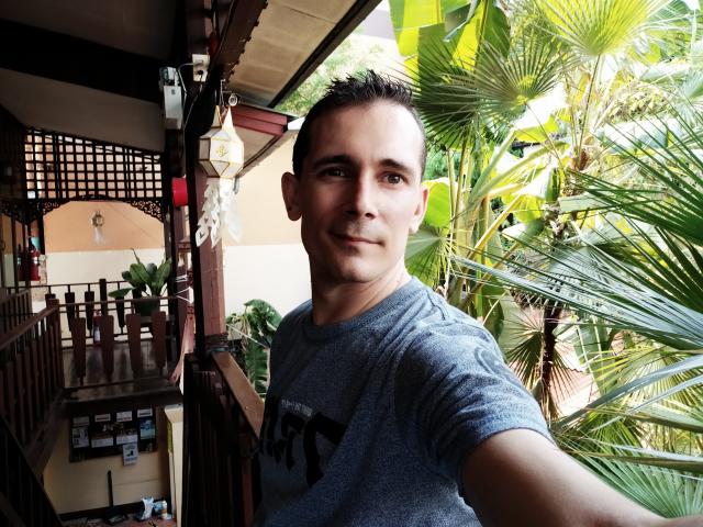 Kinkme, um rapaz atraente, de cabelo curto, com um leve sorriso, vestindo uma camiseta cinza, está em uma área tropical, de madeira e repleta de plantas, tirando uma selfie..
