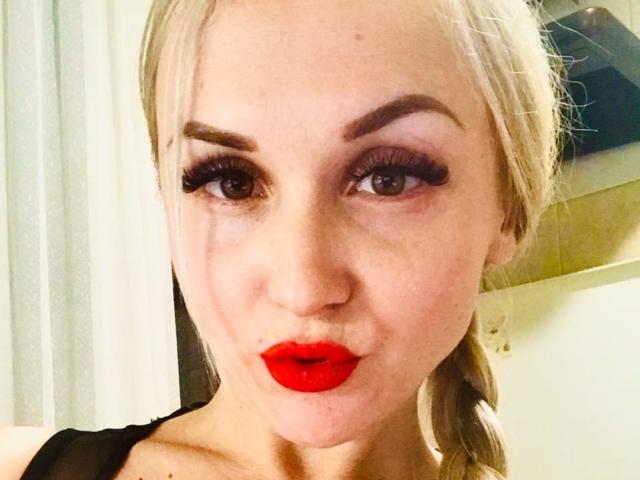 Klubnika with blonde hair, thick eyebrows, red lipstick, and heavy black eyeliner. She's making a pouty face, wearing a black top, and has large, fake-looking eyelashes.