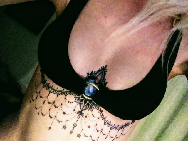 SwissMiss trägt einen schwarzen BH mit einem blauen Stein, der eine aufwendige Spitzenmuster unter ihrer Brust zeigt, mit Mustern und kleinen Punkten auf ihrer hellen Haut. Blondes Haar ist sichtbar..
