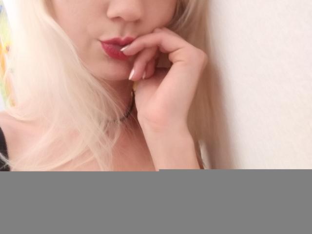 SwissMiss, avec ses cheveux blonds, son rouge à lèvres rouge et sa peau pâle, touchant ses lèvres avec sa main droite, montrant sa petite poitrine et ses ongles soignés, dans un autoportrait avec un effet de flou..