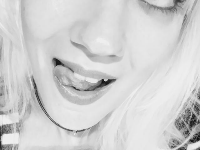 El rostro de SwissMiss, con la lengua afuera, mostrando los dientes, y los labios ligeramente entreabiertos. Tiene el cabello claro, un collar y lleva una camisa a rayas. Foto en blanco y negro..