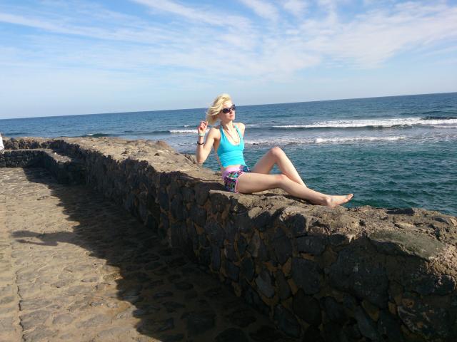SwissMiss, avec des cheveux blonds, un débardeur bleu et un bas de maillot de bain violet, assise sur un mur de pierre près de l'océan, les jambes croisées, tenant un téléphone, portant des lunettes de soleil..