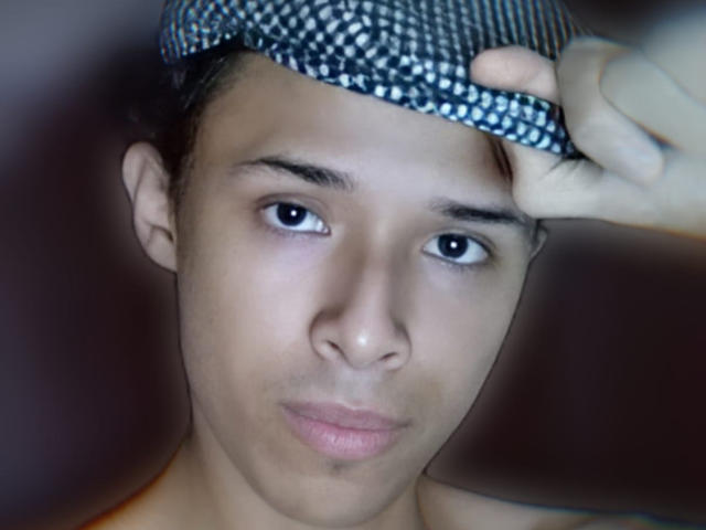 Hercolezz, coiffée d'une casquette à carreaux qu'elle tient d'une main, a une expression sérieuse, des yeux sombres et une bouche légèrement entrouverte, rosée. Son visage est lisse et elle est nue..