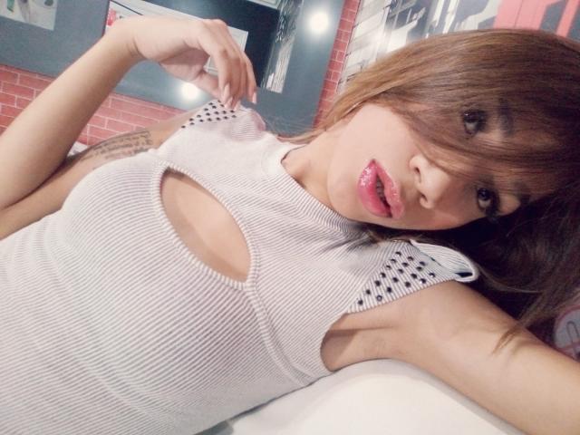Littlebrunettehot with her tits showing through a white, ribbed, cut-out top, pink glossy lips, and brown hair in a selfie, resting her arm and hand on a surface.