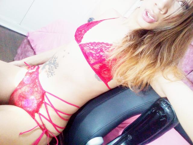 Littlebrunettehot with red lace lingerie, showing her tits and pussy, has tattoos on her side, and is sitting on a black chair with a pink pillow.