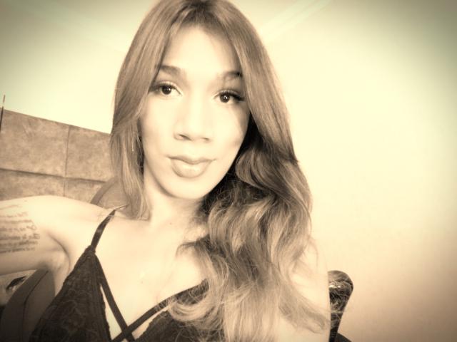 Littlebrunettehot with pouty lips, wavy hair, and a tattoo on her right arm, wearing a black lace top, in a sepia-toned selfie. Boobs visible under lace.