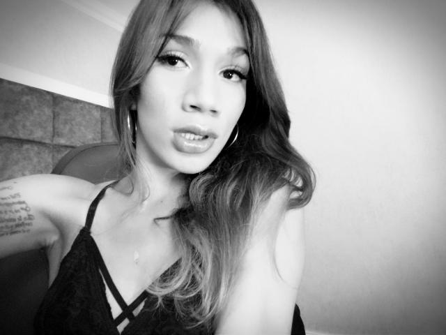 Photo en noir et blanc de Littlebrunettehot avec de longs cheveux, des boucles d'oreilles cercles, et un tatouage sur son bras droit, portant un haut en dentelle, montrant ses lèvres pulpeuses et ses petites seins..