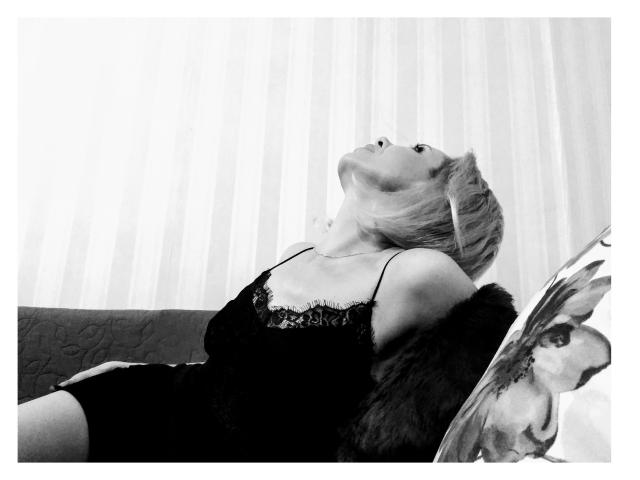Sensuellement douce, avec des cheveux blonds, une robe en dentelle noire et un manteau en fourrure, la tête penchée en arrière, les yeux fermés, détendue de manière sensuelle sur un canapé, les bras reposant. Image en noir et blanc..