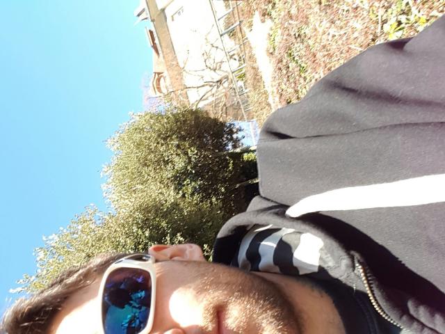 El rostro de DarkPunkX, con gafas de sol azules reflectantes, una chaqueta negra y una camisa a rayas, mirando hacia un cielo azul claro, con un arbusto y un edificio en el fondo..