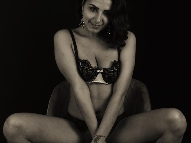 Zazazelle, com cabelo escuro, sorrindo, vestindo um sutiã de renda preto, sentada com as pernas abertas, as mãos nos joelhos, expondo seus seios e uma tatuagem no pulso direito. Fundo preto..